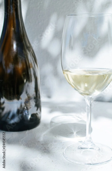 Fototapeta White wine bottle and a glass standing in front of a white concrete wall with shadows of leaves on a sunny summer day 