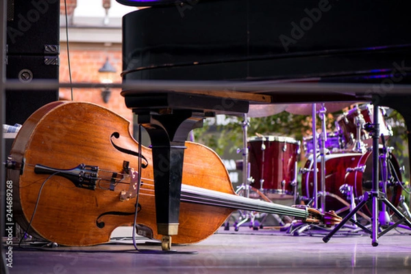 Obraz Wooden cello or violin on stage prepared for playing. Classical instruments, piano, drums in background. Concept of orchestra music. Audience waiting for concert to begin