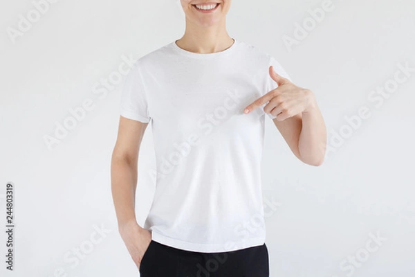 Fototapeta Young woman smiling while pointing with index finger to white t-shirt, copyspace for advertising, isolated on gray background. No face photo