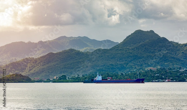 Obraz Empty Cargo ship running though in bay at Fiji country. mountains and small community are background with moody clouds during sunset.