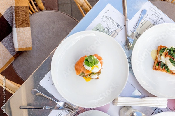 Fototapeta Breakfast table: toast with guacamole, salmon and poached egg and eggs benedict with brioche, bacon and spinach.