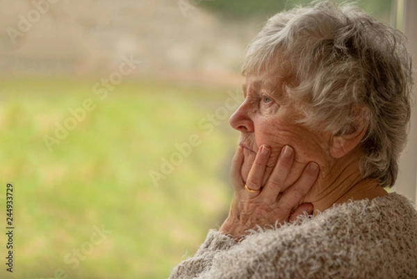 Fototapeta Senior woman looking upset and distressed through a window 