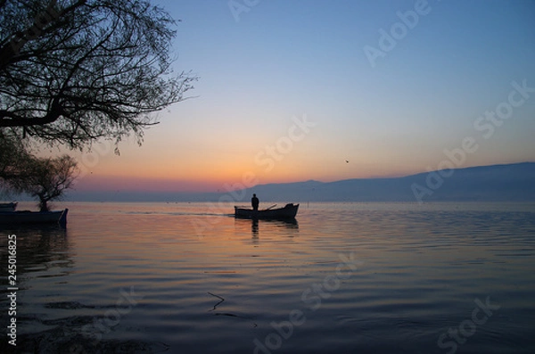 Fototapeta Golyazi, Bursa / Turkey - March 15 / 2014 : Silhouette of a fisherman staning on his boat while sunrise in the morning