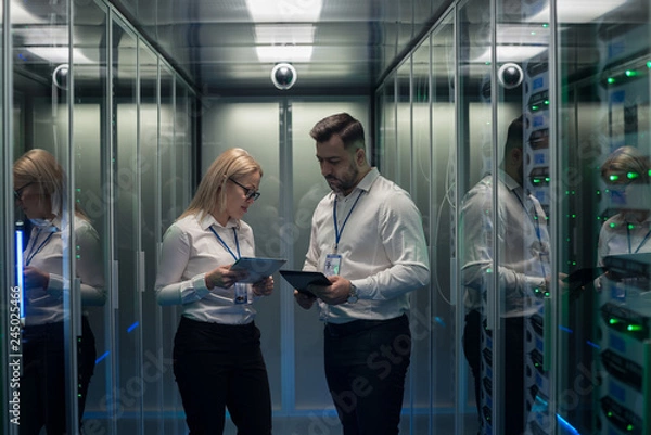 Fototapeta Side view of man and woman with tablet diagnosing server hardware opening glass door of rack in data center corridor