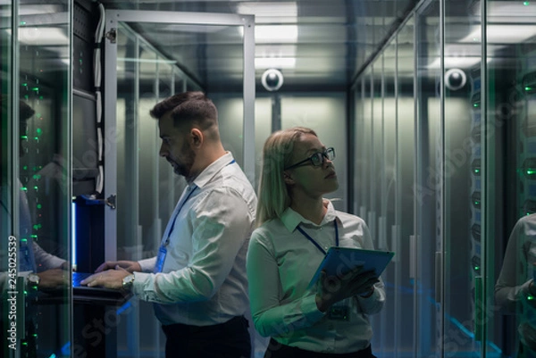 Fototapeta Side view of man and woman with tablet diagnosing server hardware opening glass door of rack in data center corridor