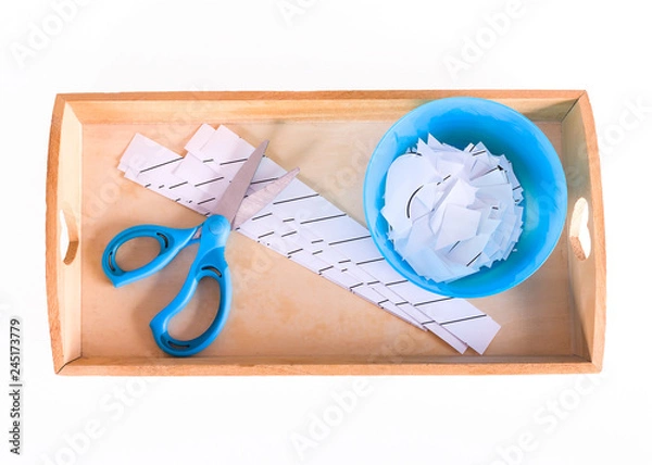 Fototapeta Children's hands hold a wooden tray with montessori materials for a lesson from the practical life zone. Blue scissors, bowl, sheets for cutting. Isolate on white background.