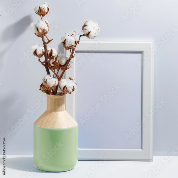 Fototapeta Mock up white frame and dry cotton twigs in blue ligth-green on book shelf or desk. Minimalistic concept.
