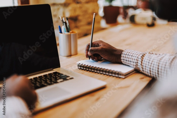 Fototapeta Close up of laptop and person making notes in notebook