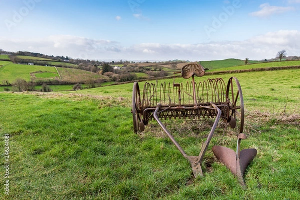 Fototapeta Old rusty horse drawn hay rake in a grass field with Devonshire countryside and hills in the background under a blue and cloudy sky on a bright day, with plenty of copy space included. Landscape.