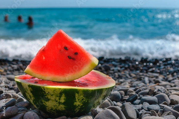 Fototapeta Rest by the sea. Sliced, juicy watermelon on pebble beach, off the coast of the blue ocean..