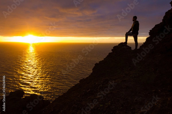 Fototapeta Hiker silhouette standing on cliff edge rock staring at sunset in Gran Canaria, Spain. Fearless man enjoys splendid twilight by the sea in Canary Islands. Visionary, adventure, challenge concepts