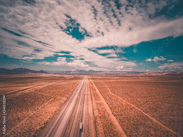 Fototapeta aerial drone shot of a highway leading off into the distance with blue skies and clouds and mountains