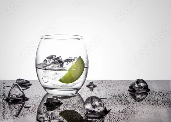 Obraz Vodka or Gin with lime in an oval glass on a glossy base with ice cubes against a white back lit background