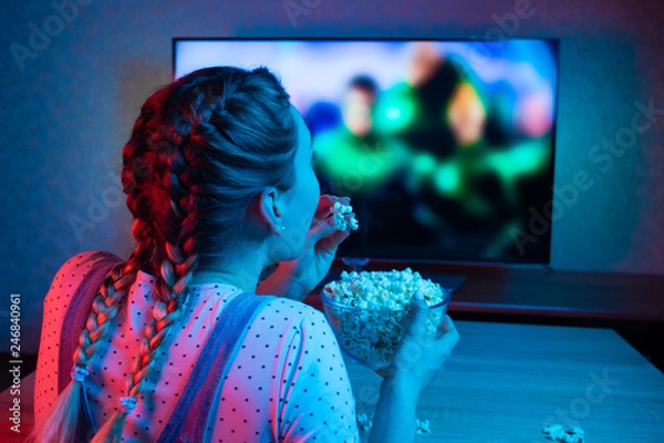Fototapeta A young girl watching movies and eating popcorn with a bowl on the background of the TV. The color bright lighting, blue and red. Relax, rest at home when watching TV, film. Background for design
