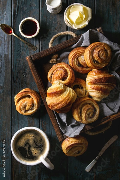 Fototapeta Variety of homemade puff pastry buns cinnamon rolls and croissant served with coffee cup, jam, butter as breakfast over dark plank wooden background. Flat lay, space