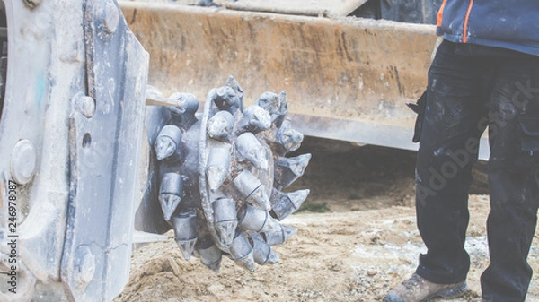 Fototapeta Ein Arbeiter steht neben dem Bohreraufsatz eines Baggers, mit dem Stein und Fels abgetragen werden kann.  