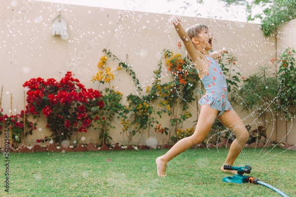 Obraz Funny laughing little girl  running though garden sprinkler playing with water splashes  in the backyard on a sunny hot summer vacation day .