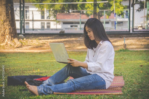 Fototapeta The asia woman is playing computer on a notebook.She sits and play laptop in park.