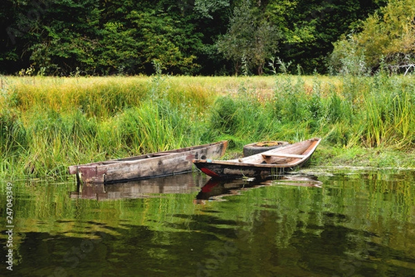 Fototapeta Two old boats on the shore are tied to an old car wheel, on a background of green grass a sunny summer day