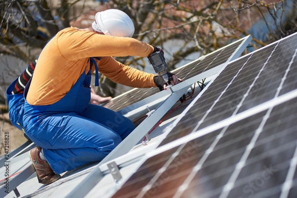Fototapeta Male worker in protective helmet installing stand-alone solar photovoltaic panel system using screwdriver. Electrician mounting blue solar module on roof of modern house. Alternative energy concept.