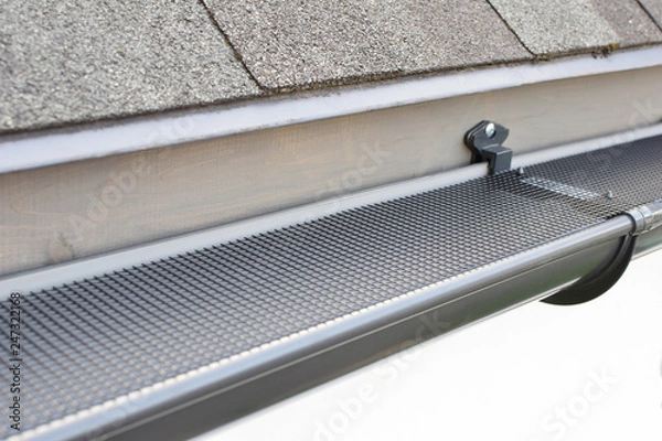 Obraz Plastic guard over new dark grey plastic rain gutter on asphalt shingles roof at shallow depth of field.