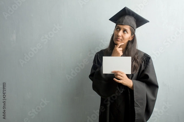 Fototapeta Young graduated indian woman against a wall thinking and looking up, confused about an idea, would be trying to find a solution. Holding a placard.