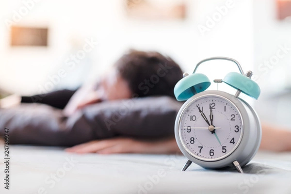 Fototapeta Alarm clock in the morning. Young man sleeps in the blurry background.