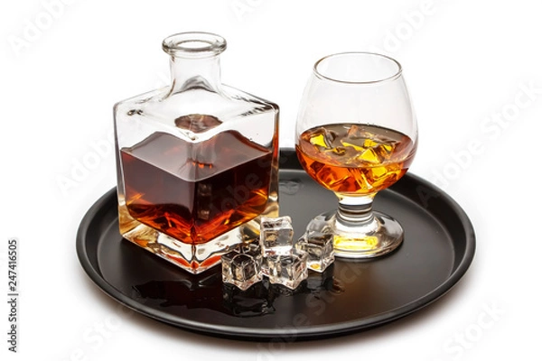 Fototapeta A glass of whiskey with ice or brandy and a square carafe on a black dish isolated against a white background. Whiskey with ice in the glass on a white background.
