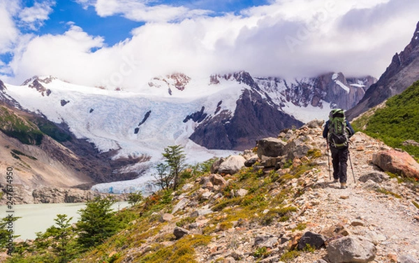 Fototapeta Young man walking up to mount Torre view point next to a lake or lagoon, with a green backpack on, on a summer sunny day with mountains and a glacier, near campsite De Agostini, El Chalten, Patagonia