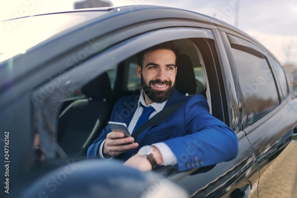 Fototapeta Close up of smiling businessman using smart phone while sitting in his car and leaning on the window.