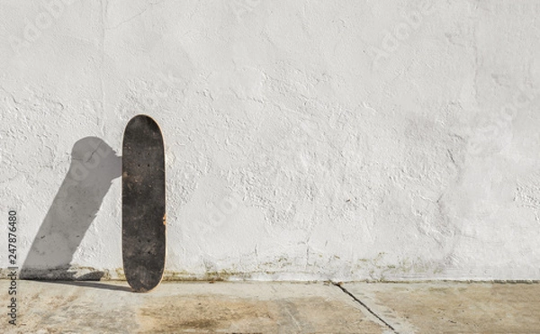 Obraz Skateboard on deteriorated white wall during a sunny afternoon with big copy space. Skateboarding background, street sport.