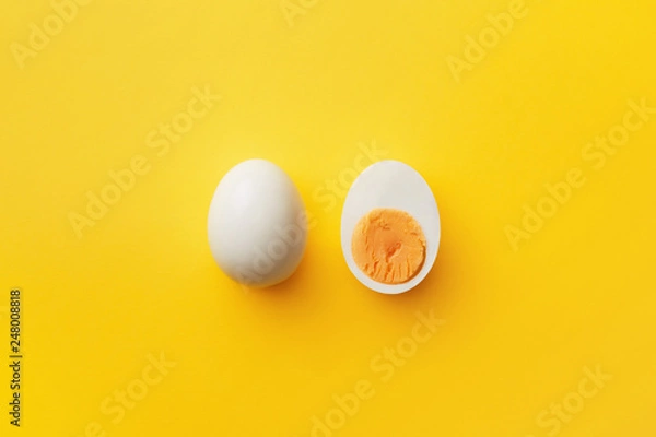 Fototapeta Single whole white egg and halved boiled egg with yolk on a yellow background. Top view
