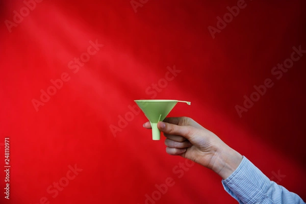 Obraz Female hand holding a green funnel on a red background, rays of light. Sifting the truth. Be able to analyze.