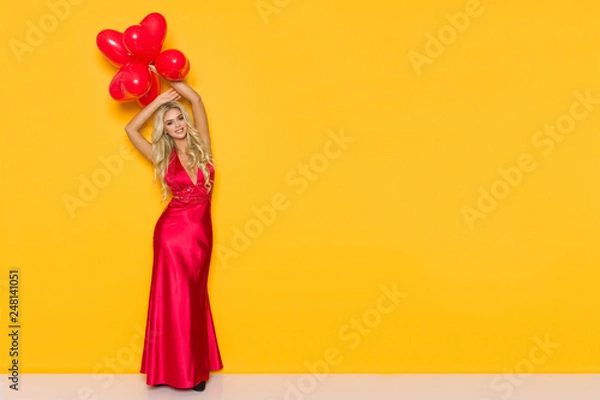 Fototapeta Smiling Beautiful Woman In Elegant Red Dress Is Holding Heart Shaped Balloons Over Her Head