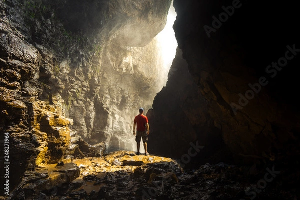 Obraz Portrait of a Male Backpacker wearing a red shirt standing in sunlight in a cave beneath the Elephant waterfall in Dalat Vietnam.