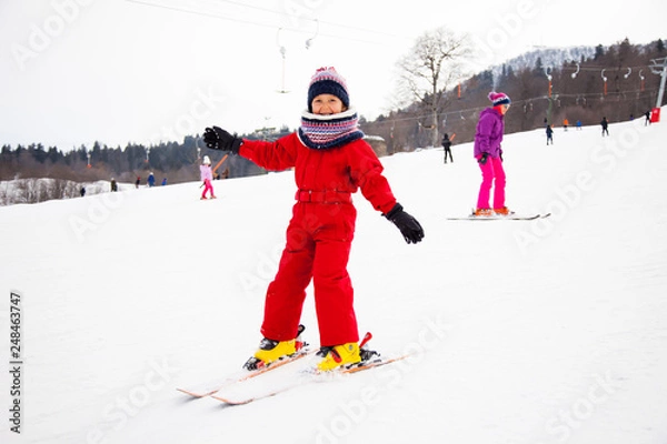 Fototapeta Little smiling girl in red overall training skiing