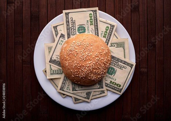 Obraz Conceptual photo. Hamburger with dollars on a white plate and on a wooden background. Top view..