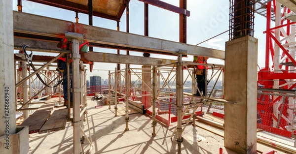 Fototapeta high building under construction with workers in the construction site on supports wearing safety uniform