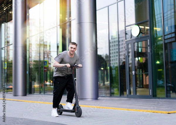 Obraz Attractive young man riding a kick scooter at cityscape background.