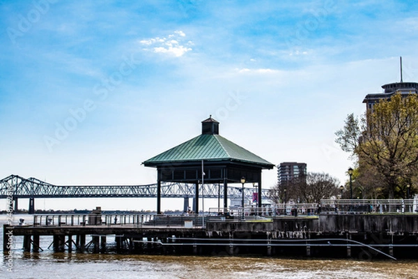 Fototapeta Buildings and a Green Gazebo Line the Waterfront with the Crescent City Connection Bridge in the Background along the Mississippi River in New Orleans, Louisiana, USA