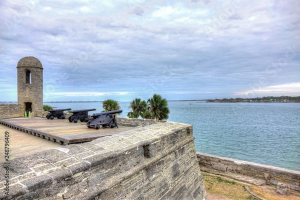 Fototapeta Three Spanish colonial cannons mounted on top of an old Spanish fort, overseeing the ocean, a cloudy sky and palm trees. Shot in Castillo de San Marcos, St Augustine, Florida, USA.