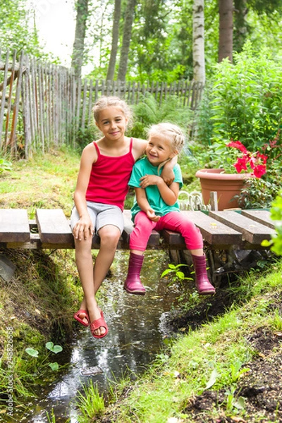 Obraz Happy smiling children sitting on the bridge at the creek embracing one another in a country house garden. Kids playing outdoors in summer, vertical