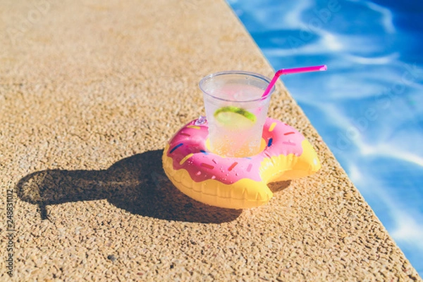 Fototapeta Tasty refreshing cocktail on on inflatable donut plastic toy on edge of swimming pool. Ripple Water in swimming pool with sunny reflection
