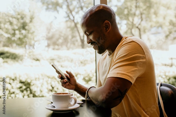 Fototapeta Happy cheerful African hipster holding mug, drinking fresh cappuccino, browsing internet and checking newsfeed on social media.Man using cell phone during coffee break at modern cafe