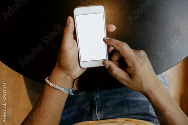 Fototapeta Closeup view of male hands touching mobile phone.African man using smartphone while sitting at the wooden table.Empty white blank copy space screen for your information.Mockup.
