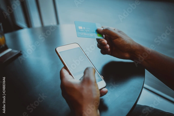 Fototapeta Closeup view of male hands touching mobile phone.African man using smartphone and credit card while sitting at the wooden table.Empty white blank copy space screen for your information.Mockup