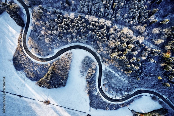Fototapeta Aerial drone view of a curved winding road through the forest up in the mountains in the winter with snow covered trees and curved streets in winter while sunset and sun rays casting trough