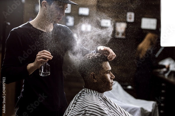 Fototapeta The barber dressed in a black clothes is doing the hair styling to the stylish black-haired  man sitting in the armchair in a barbershop