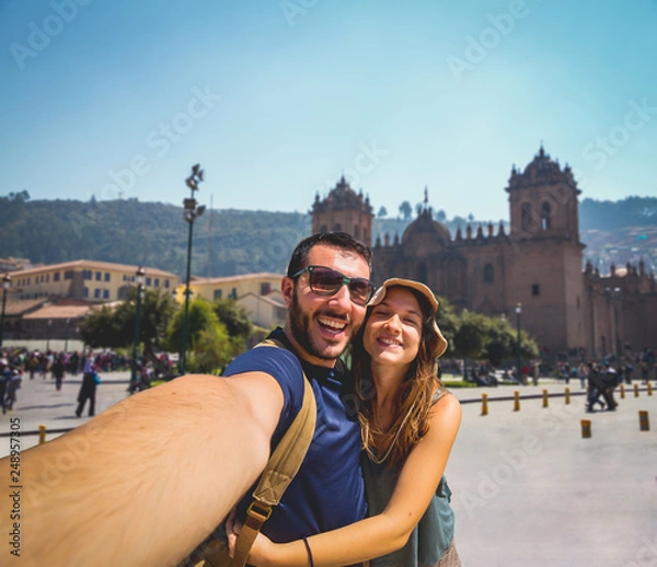 Obraz handsome happy tourist couple taking selfie in Plaza de Armas with Inca fountain, Cathedral and Compania de Jesus Church, city of Cusco, Peru