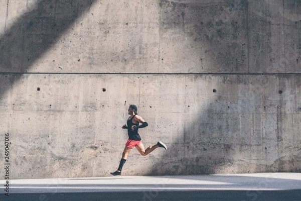 Fototapeta Muscular Male athlete sprinter running fast,exercising outdoors,jogging outside against gray concret background with copy space area for text message or ad content.Side view,full length